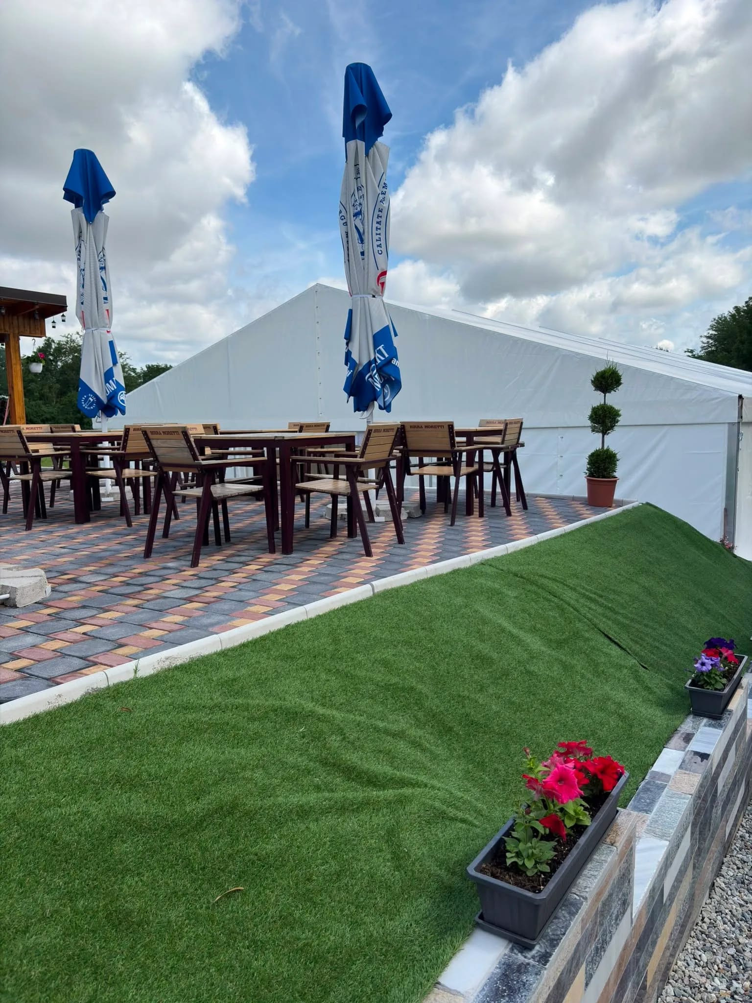 Mese și scaune de lemn aranjate pe terasa pavată, lângă peretele cortului, cu jardiniere cu flori pe margine.