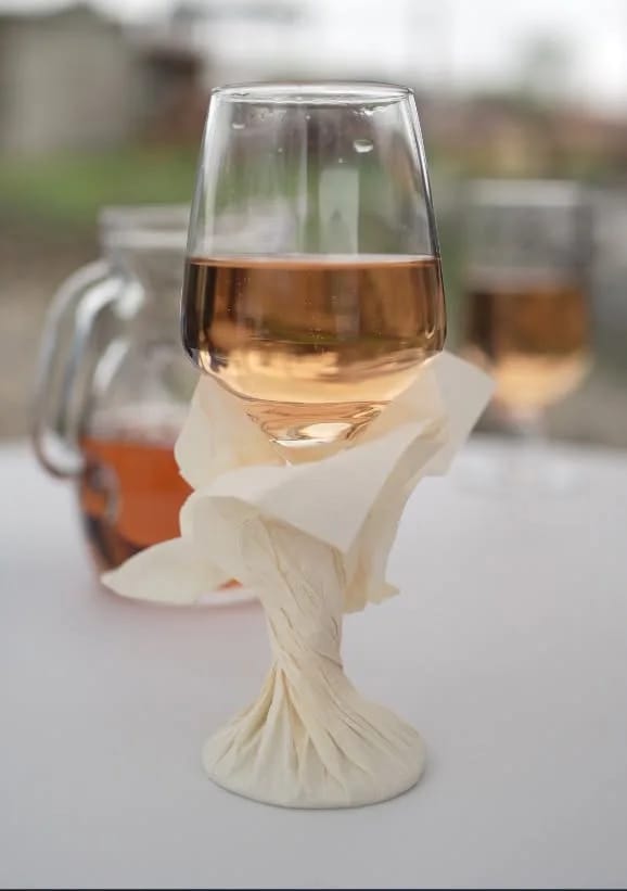 Pahar de vin rosé pe o masă îmbrăcată în alb, cu șervetul pliat în jurul piciorului și o carafă în fundal.
